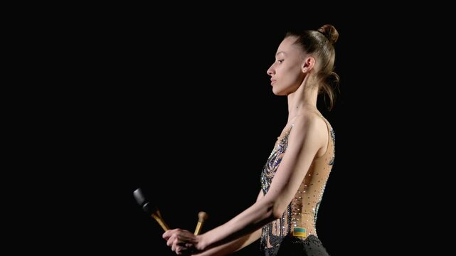 Sie view of professional pretty rhythmic gymnast in elegant leotard practicing rhythmic gymnastic elements, performing small throw and catch with clubs over black background.