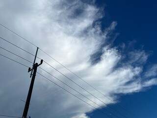 This photo captures a dramatic sky with a mix of wispy white clouds and deep blue background, creating a dynamic contrast. In the foreground, a utility pole with three visible power lines s