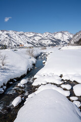 A clear mountain stream flows between snow-covered banks, surrounded by frosty trees and snowy peaks under a bright blue sky. A peaceful winter landscape showcasing nature's pure, icy beauty.