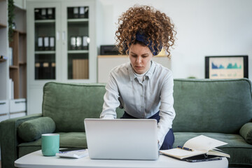Confident Russian businesswoman with curly hair sits on modern office sofa, writing in notebook. plans strategies and documents ideas with thoughtful intent and professional poise.