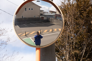 Photographer taking a self-portrait through a convex traffic mirror on a quiet street, capturing reflection on a sunny day. Creative perspective, urban exploration, and street photography concept.