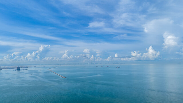 Scenic aerial view of a peaceful seascape with a long pier extending into calm blue waters, surrounded by ships and distant skyline under a vast cloudy sky. Maritime and coastal development concept.