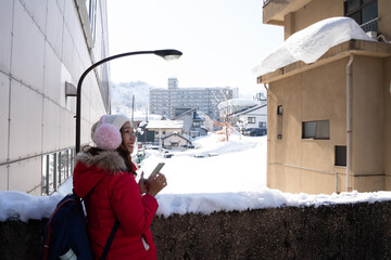 Smiling woman in red winter coat and pink earmuffs using smartphone in a snowy city street. Bright sunny day, cheerful travel moment, and winter lifestyle in an urban environment.
