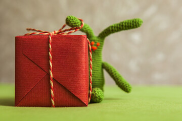Close up of red wrapped gift box with braided rope bow and partially visible green crochet bunny in...