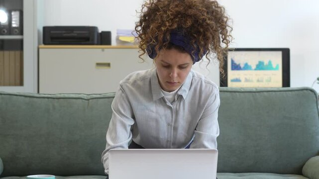 Confident Russian businesswoman with curly hair sits on modern office sofa, writing in notebook. plans strategies and documents ideas with thoughtful intent and professional poise.