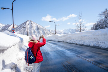 A person in red winter clothes walks along a snow-covered mountain road under a clear blue sky....