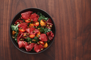 top view of salad with kale, bresaola and cherry tomatoes decorated with capers in black bowl on walnut wood table