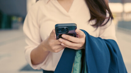 Close-up of Asian businesswoman holding smartphone in both hands, with jacket draped over arm....