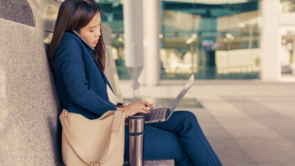 Asian businesswoman multitasking with phone and laptop while sitting outdoors. Working on...