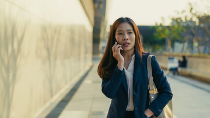 Asian businesswoman in formal suit talking on phone while walking along modern urban sidewalk....