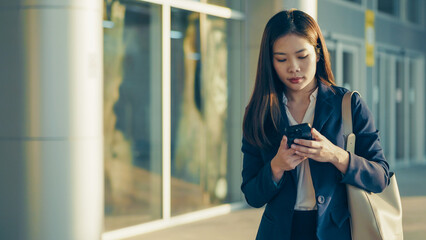 Asian businesswoman in formal suit using smartphone while walking in modern city area. Confident and focused expression in outdoor urban environment.