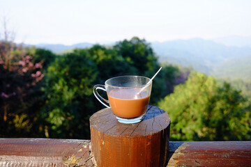 A glass cup of hot coffee on wooden fence with the valley mountain in natural sunlight of morning...
