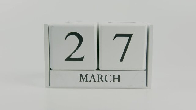 A hand flips a wooden calendar from March 27th to March 28th on a white background, marking the passing of time and the start of a new day