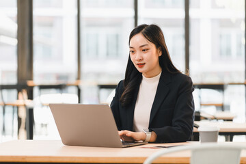Fototapeta premium Young Asian businesswoman in black suit working on laptop at modern office, focused and professional, natural light from large windows, coffee cup on desk, productivity concept