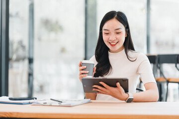 Young Asian woman smiling while working on tablet at modern office desk, holding coffee cup, feeling happy and productive, with documents and pen on table