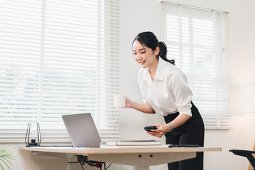 Young Asian woman in white shirt standing at modern office desk, holding coffee cup and smartphone, smiling while looking at laptop, bright window background