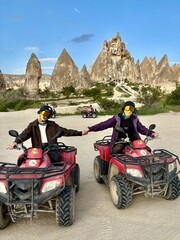 Two friends ride ATVs in the unique landscape of Goreme, Cappadocia. With hands joined, they explore towering rock formations under a bright sky, sharing an exciting outdoor adventure in Turkey