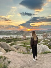 A girl stands atop a rock formation in Cappadocia, Turkey, gazing at the breathtaking sunset over the valleys and unique rock formations. The colors of the sky create a serene atmosphere