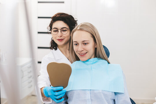 Young dentist and her young female patient smiling while looking in the mirror in dental clinic