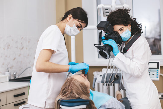 Female dentist with camera making shots of patient's smile after treatment using special shadowless camera with flash ring. Medical assistant holding mirror.