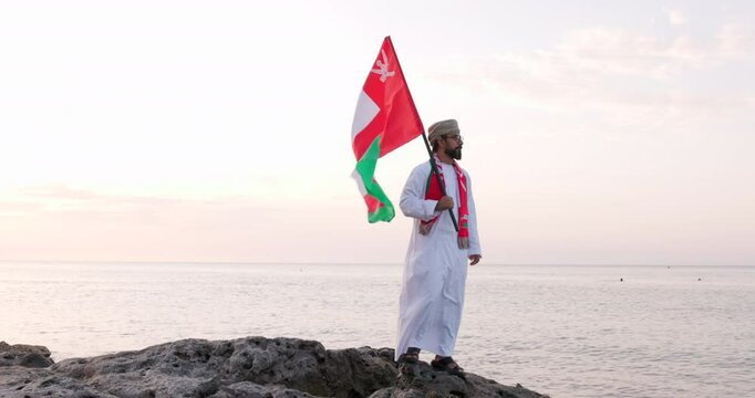 Omani man stands with Omani National Flag, wearing omani traditional dishdasha and Musaar