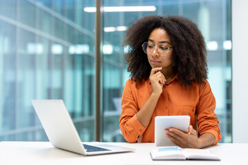 A thoughtful businesswoman in glasses looks at a tablet and considers her next business move. The environment appears to be a modern office with large windows.