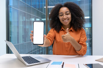 A cheerful young woman with glasses shows a blank smartphone screen and gives a thumbs up in her office setting.