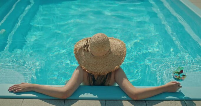 Young woman relaxing in a swimming pool with a cocktail, wearing a straw hat and enjoying a sunny summer day
