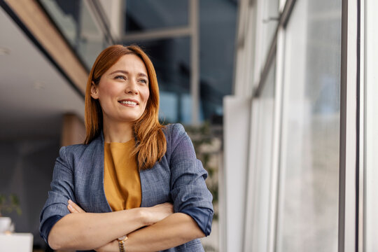 Portrait of confident professional businesswoman standing at corporate firm with arms crossed and looking through the window.