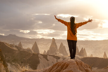 Back view of a tourist girl with her arms raised to the sides on the edge of a cliff in Cappadocia, Turkey. Beautiful sunset landscape