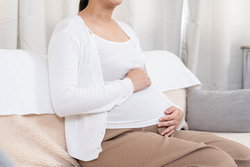 Closeup of pregnant Asian woman gently placing hands over round belly sitting on soft sofa wearing white cardigan and ribbed pants showing peaceful moment of maternity in cozy home setting