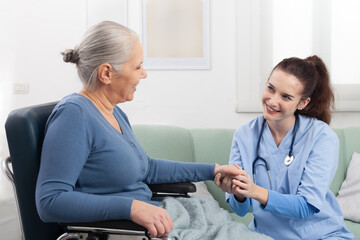 Fototapeta premium Smiling young nurse assisting an elderly woman in a wheelchair at a healthcare or nursing home facility. Concept of medical senior health care, geriatric assistance, and quality of life in old age.
