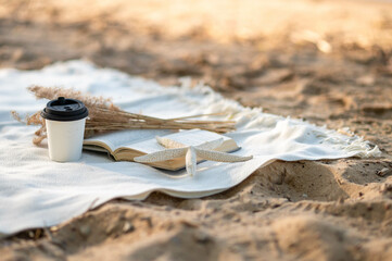 Open book, dried flowers, hot drink, and starfish on a blanket spread on yellow sand in a cozy boho scene.