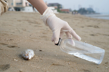 A person in gloves picks up a bottle of water near a dead fish on the beach, highlighting environmental concerns about pollution and marine life.