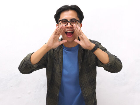 Excited young Asian man in a blue t-shirt making an announcement gesture,young Asian man wearing blue t-shirt doing whisper gesture  against a white background.
