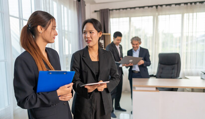 Two businesswomen discussing project details using tablet and clipboard during workday in office, collaborating on ideas, showing teamwork and communication, colleagues in background reviewing laptop
