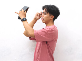 A young Asian man in a pink shirt holds an empty wallet with a sad expression on a white isolated background.