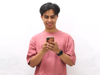 Young Asian man in a pink T-shirt smiling while holding and focusing on a mobile phone against a white background.
