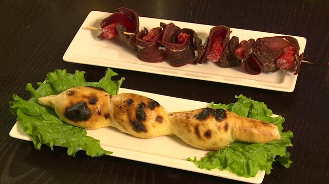 Appetizing cured meat skewers and uniquely twisted baked bread on white plates. Traditional restaurant starters or savory snacks presented on a dark wooden table for culinary concepts.