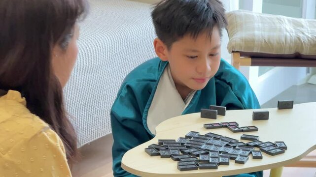 The Southeast Asian boy is playing a domino game with his mother in the living room.