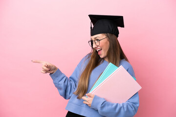 Young student woman wearing a graduate hat isolated on pink background pointing finger to the side...