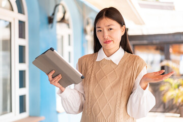 Young Chinese woman holding a tablet at outdoors making doubts gesture while lifting the shoulders