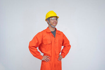 Young Southeast Asian male construction worker wearing safety helmet and goggles, confidently standing with hands on hips in a dependable pose. Isolated on a light background.