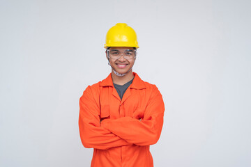 Young Southeast Asian male construction worker smiling with arms confidently crossed, wearing safety gear and orange coveralls. Isolated on a light background.