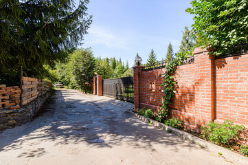 driveway leads through a gate with brick pillars into a lush, green property on a sunny day. A wooden fence runs alongside the road