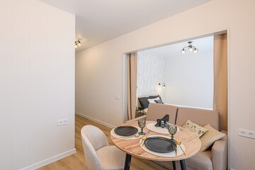 Cozy apartment interior featuring a dining area with a round table, chairs, and a sofa, leading to a bedroom. Neutral tones dominate.