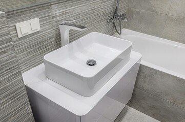 Modern bathroom scene featuring a rectangular white sink with a sleek faucet, set on a white cabinet against gray tiled walls. A bathtub is visible in the background