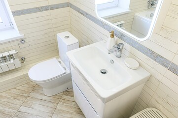 Bright bathroom with white fixtures, including a toilet, sink, vanity, and mirror. The walls and floor are tiled with a wood-like texture