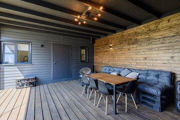 Outdoor dining area with wooden deck, table, chairs, and pallet sofa. Rustic wall contrasts with the grey siding.