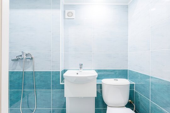 clean bathroom features light and teal-blue tiles, a white sink with a cabinet, a toilet, and a showerhead. A ventilation fan is on the wall.